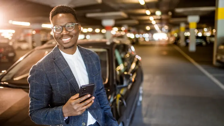 A smiling man in a suit and glasses leans against a car in a well-lit parking garage, holding a smartphone in his hand. The background features parked cars and ceiling lights.